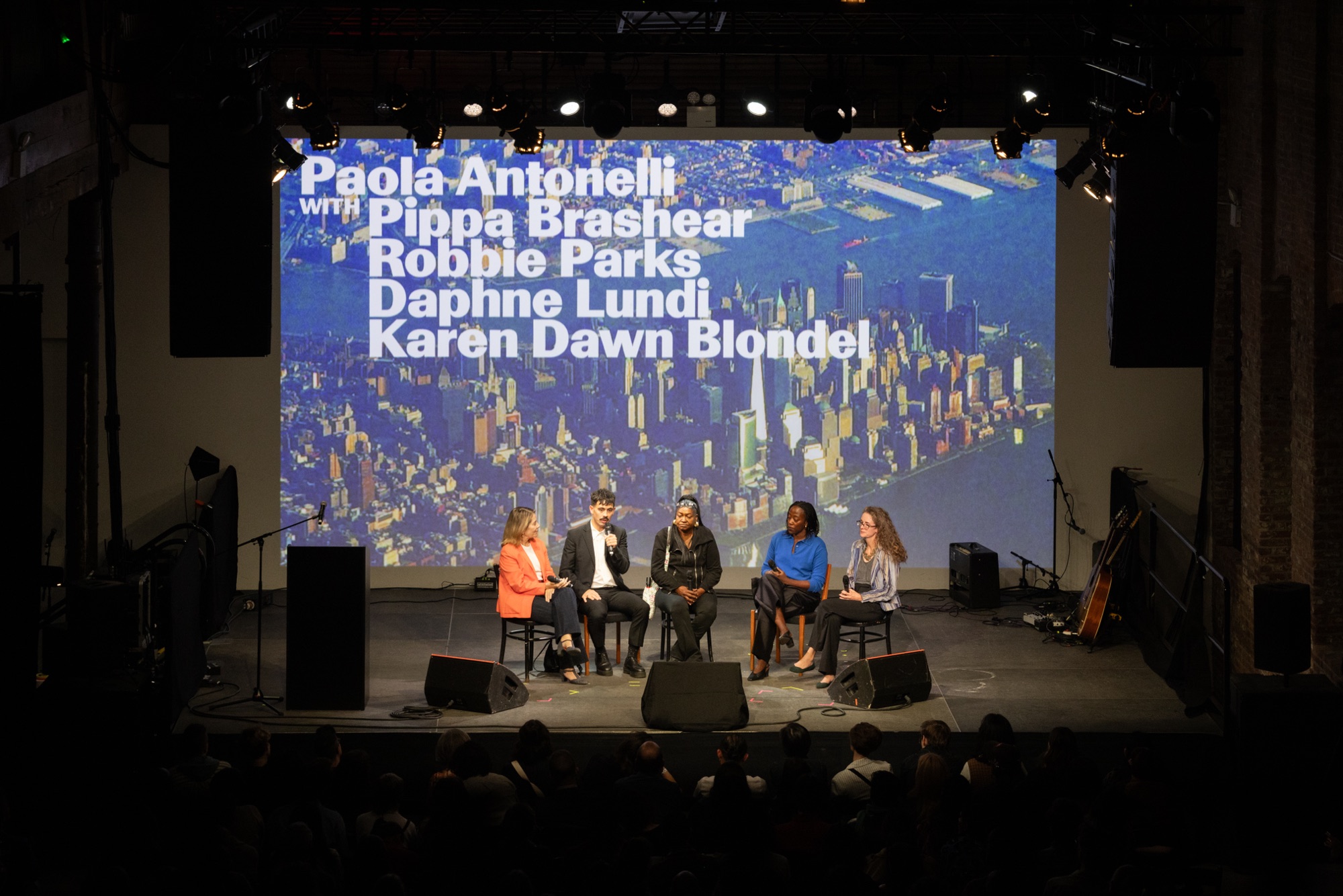 Paola Antonelli in conversation with Pippa Brashear, Robbie Parks, Daphne Lundi, and Karen Dawn Blondel on the Pioneer Works stage, an aerial image of lower Manhattan projected behind them